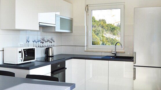 A modern kitchen is equipped with essential appliances, including a microwave and a kettle. Sleek white cabinets are complemented by a dark countertop. A large window allows natural light to fill the space, offering views of the surrounding greenery.