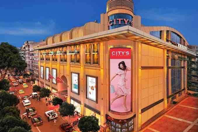 A large shopping center is depicted, featuring a modern architectural design with a bright lit facade. Multiple storefronts showcase large advertisements and signage. Surrounding streets are filled with cars, and trees line the sidewalks, contributing to a vibrant urban atmosphere.