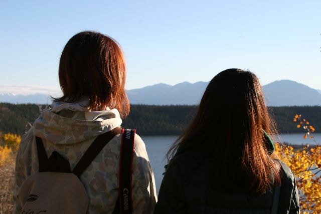 Two individuals are seen from behind, gazing out over a serene lake surrounded by mountains. The clear sky highlights the natural beauty, and autumn foliage can be observed in the foreground, enhancing the peaceful setting.