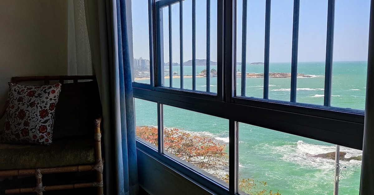 A large window frames a stunning view of the ocean, with rolling waves and a glimpse of distant coastline. Natural light floods the space, highlighting the nearby seating area adorned with a decorative cushion.