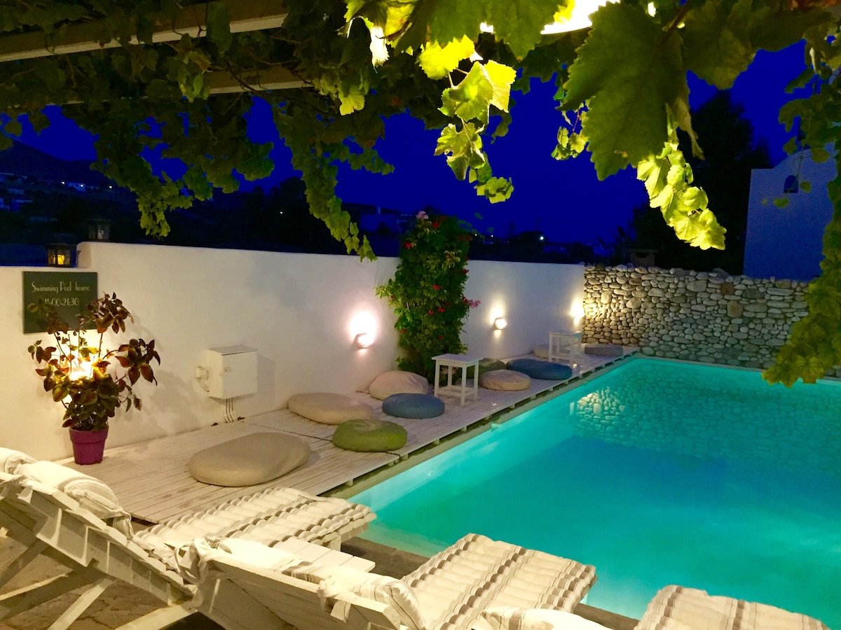 An outdoor area is presented with a well-maintained pool, surrounded by comfortable lounge chairs and colorful cushions. Green vines drape from the overhead beams, enhancing the serene setting lit by soft wall sconces. The tranquil water reflects the evening sky, creating a peaceful ambiance.
