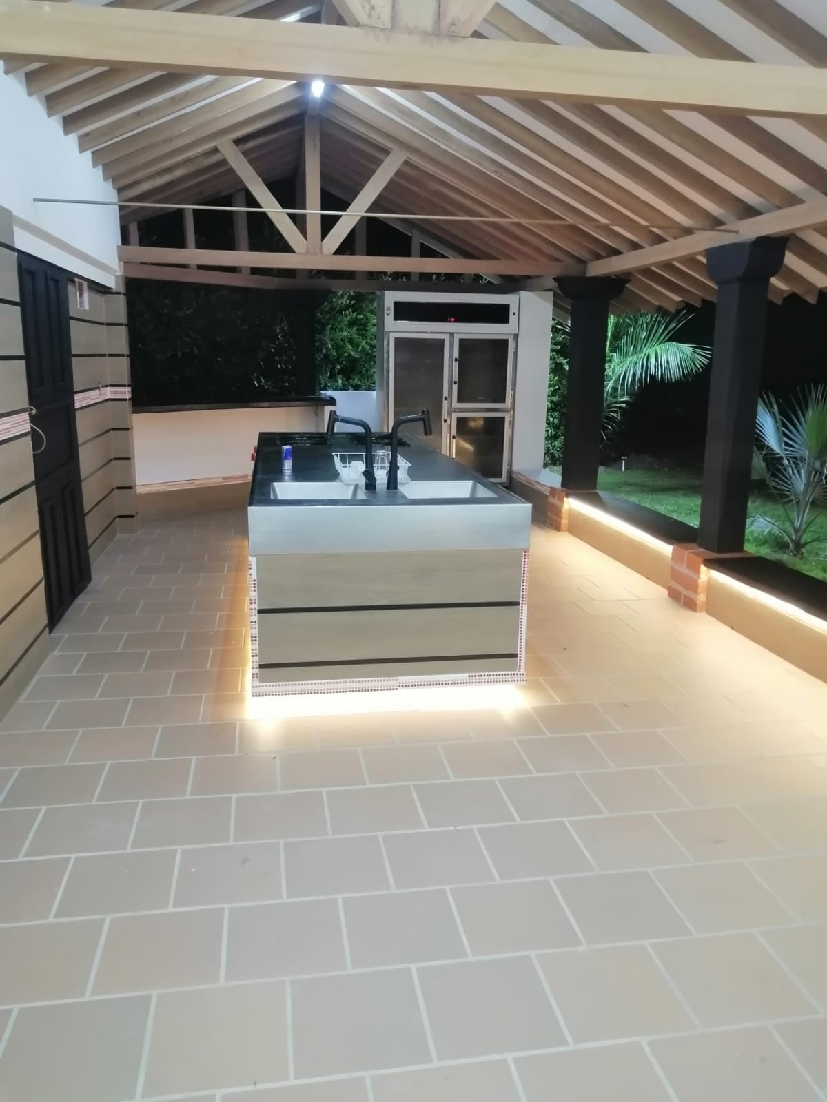 An outdoor kitchen area is featured under a wooden roof with exposed beams. A sleek kitchen island with a dark countertop is prominent, illuminated by recessed lighting along the base. The surrounding space is accented by green foliage, highlighting the tranquil outdoor setting.
