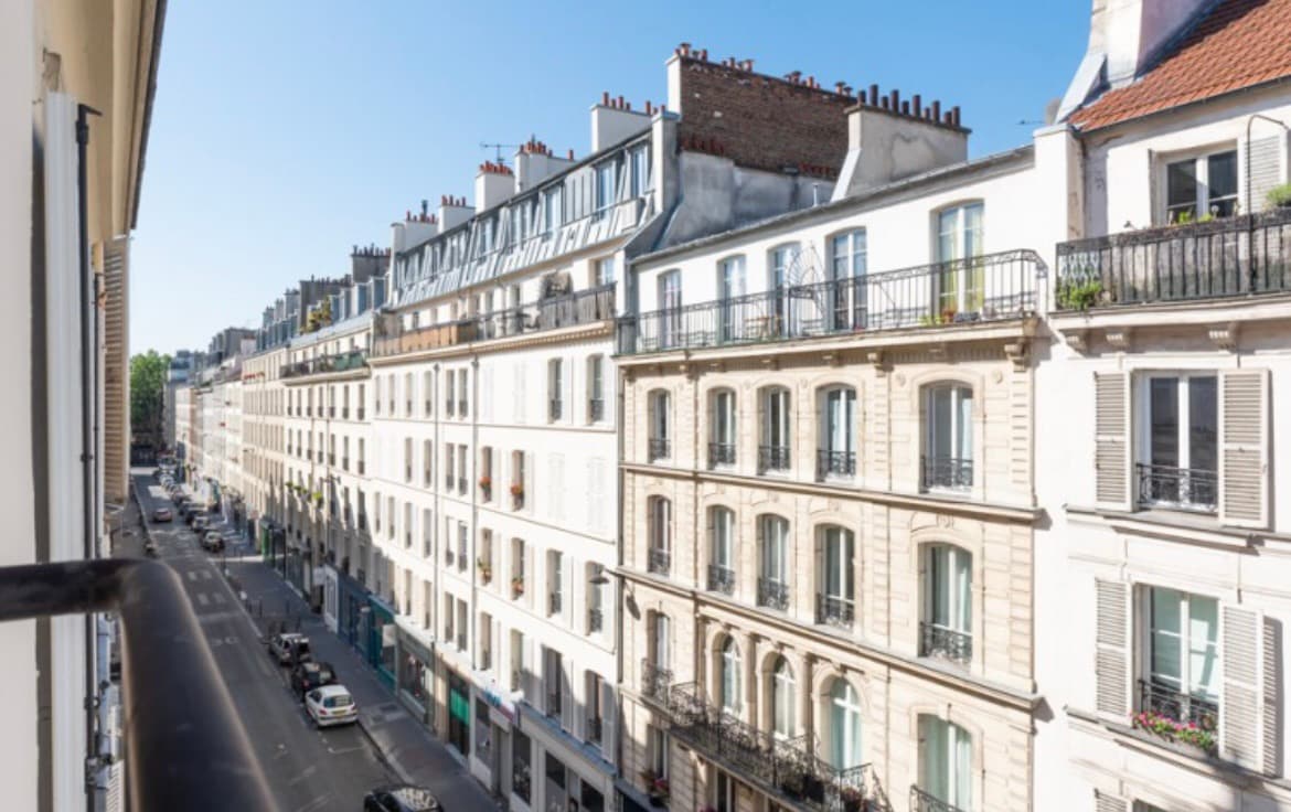 A view of a tree-lined street in a vibrant neighborhood, showcasing a mix of historical buildings with balconies. The sky is clear, and sunlight illuminates the façades, enhancing the architecture. Cars line the street below, suggesting an active urban atmosphere.
