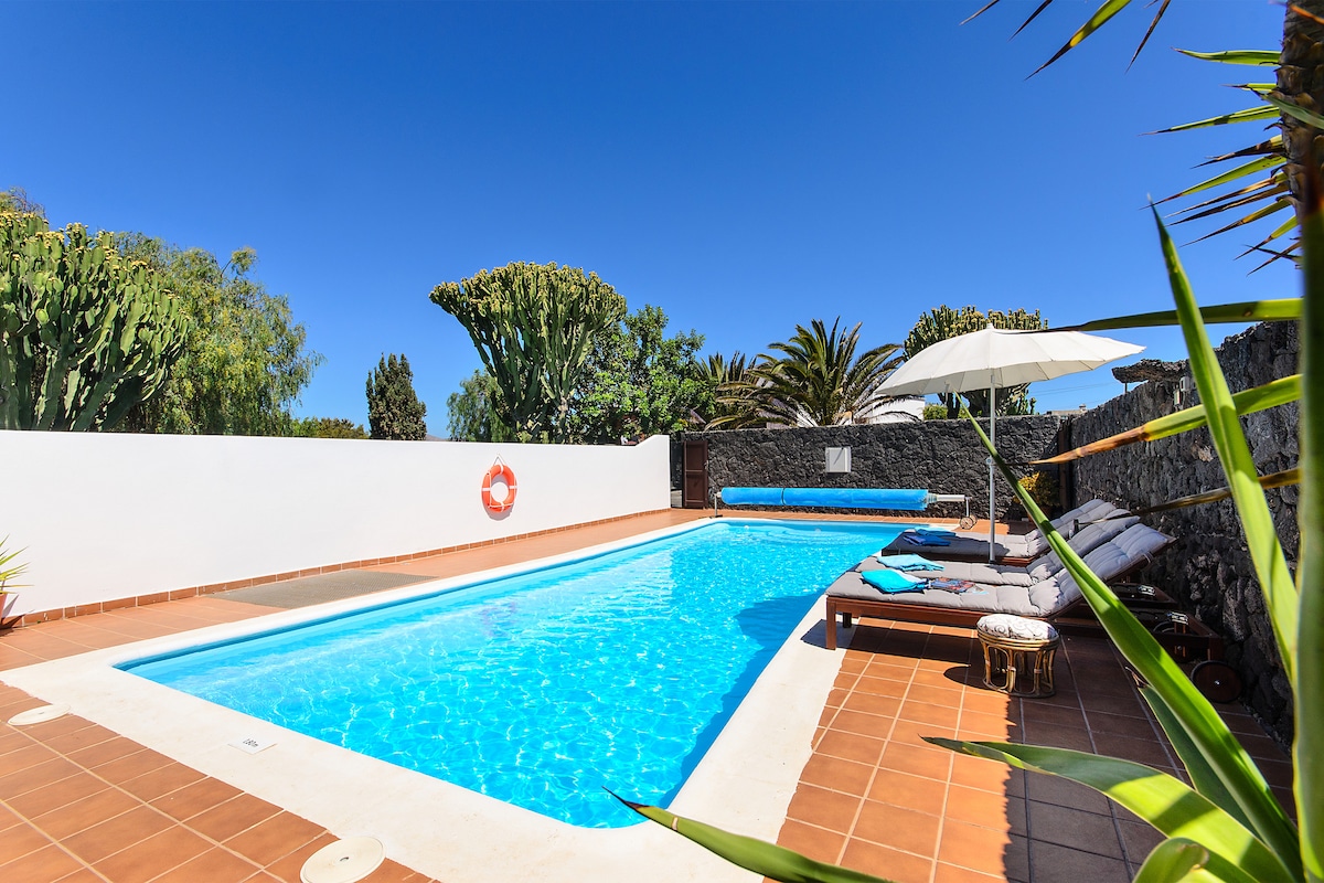 A private swimming pool is surrounded by a sun terrace featuring comfortable loungers and an umbrella for shade. Lush greenery and cacti are visible in the background, with a clear blue sky above, creating an inviting outdoor space.