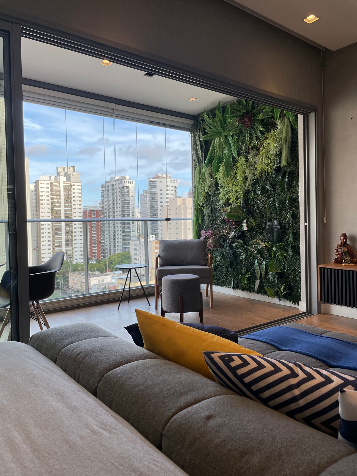 A living area featuring a comfortable gray sofa positioned near large glass doors that open to a balcony. A vertical garden with lush greenery adds a natural touch to the space, while a modern chair and colorful cushions enhance the inviting atmosphere.