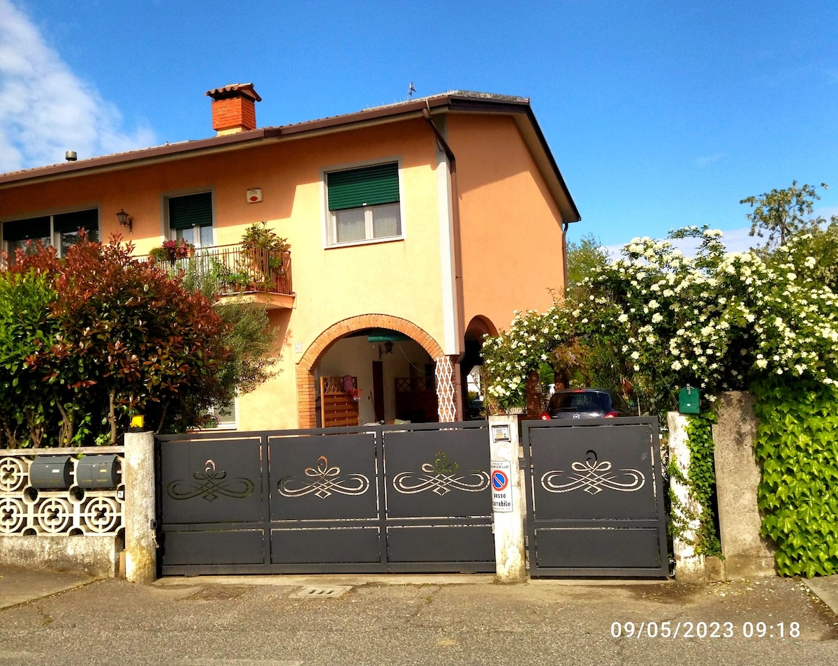 A charming two-story residence is highlighted, with a soft-colored façade and green shutters. A wrought-iron gate provides access to the property, framed by lush greenery and flowering plants. The entrance features a welcoming archway, suggesting a warm reception for guests.