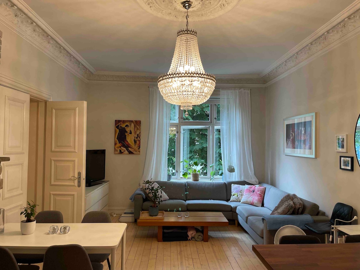 A well-lit living room features a large sofa adorned with colorful pillows, paired with a wooden coffee table. A dining table with six chairs is visible, along with a decorative chandelier hanging from the ceiling. Large windows allow natural light to enter, framed by sheer curtains.