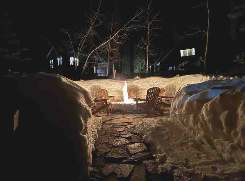 A firepit area is surrounded by snow-covered terrain, creating a cozy gathering space at night. Adirondack chairs are arranged around the flames, offering a spot for warmth and relaxation. Soft lighting from nearby homes adds a serene ambiance to the winter landscape.
