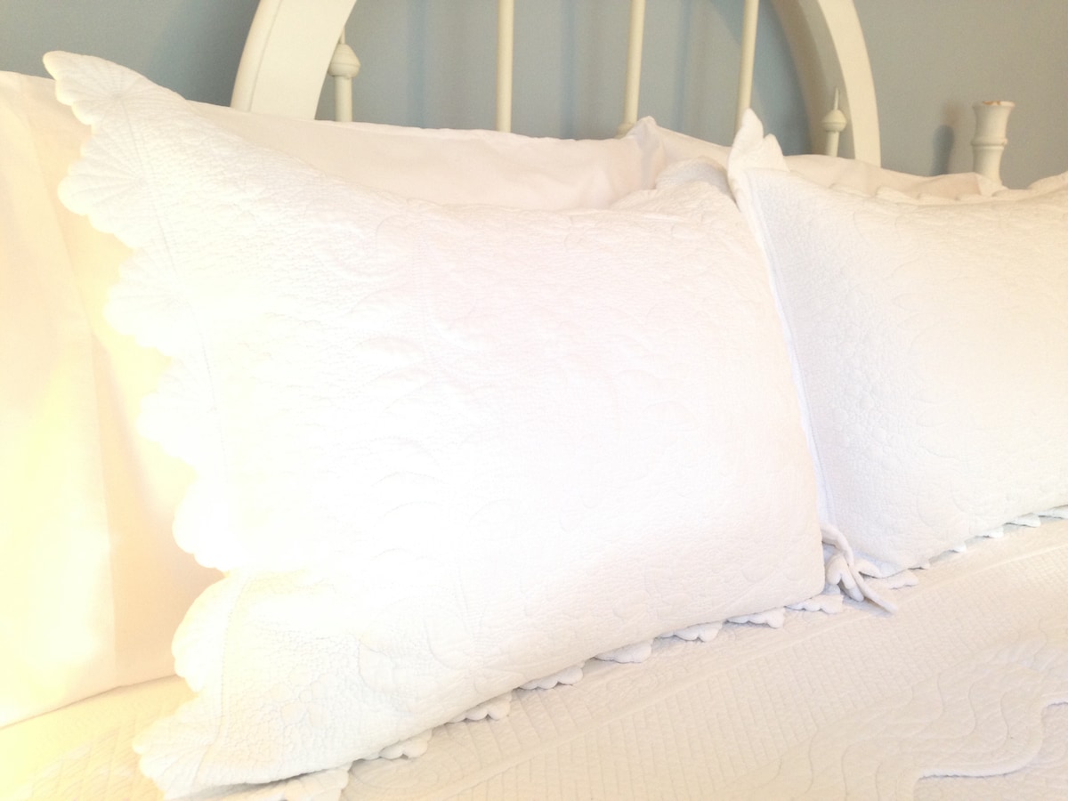 Two white quilted pillows are positioned on a bed, accompanied by smooth white linens. A scalloped edge decorates the pillows, enhancing their textural detail against the soft, light backdrop.