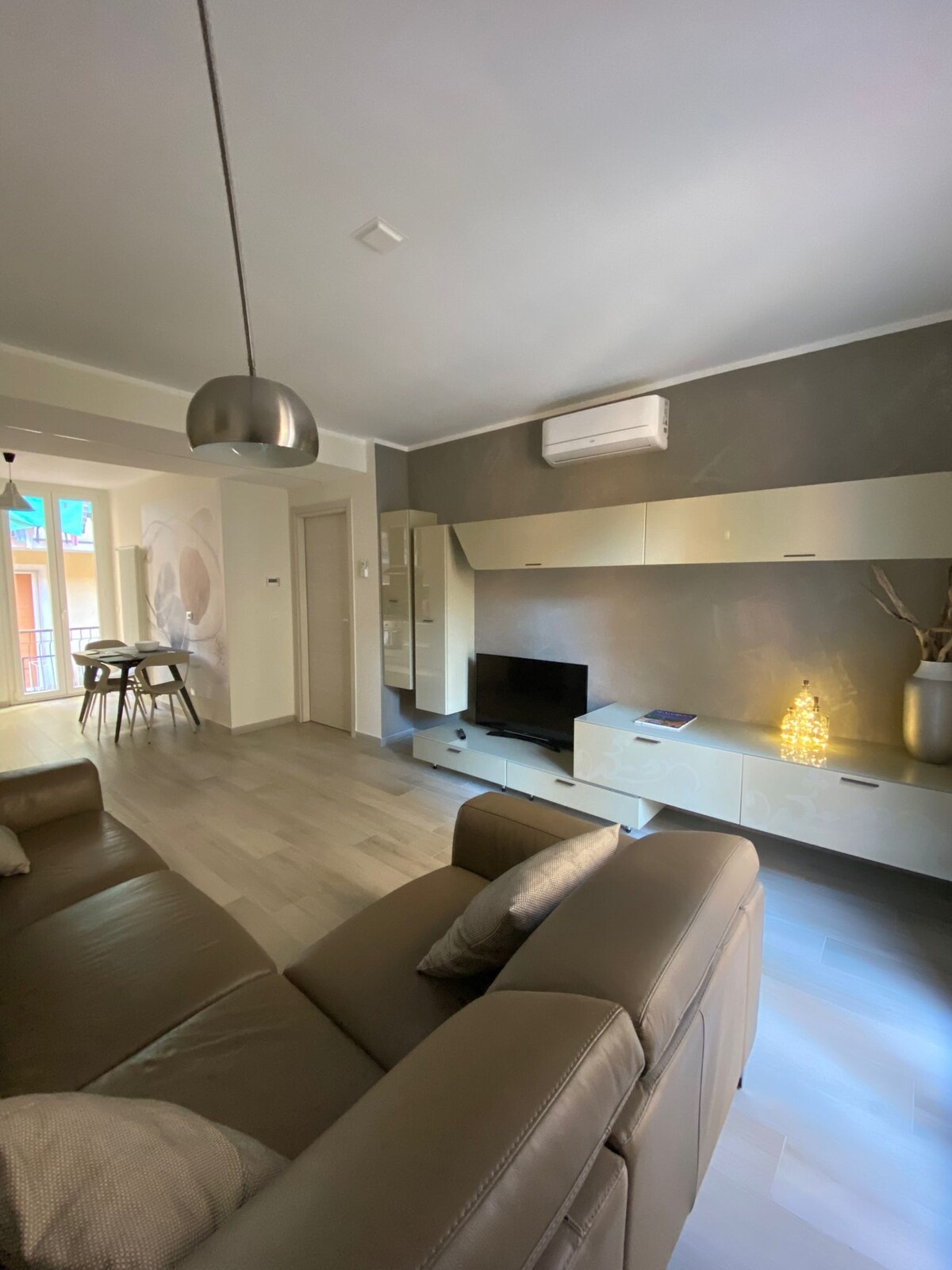 A modern living area is presented with a comfortable leather sofa and a sleek entertainment unit. Natural light flows in from a nearby window, and air conditioning is visible on the wall. A small dining table is situated in the corner, complementing the neutral color scheme.