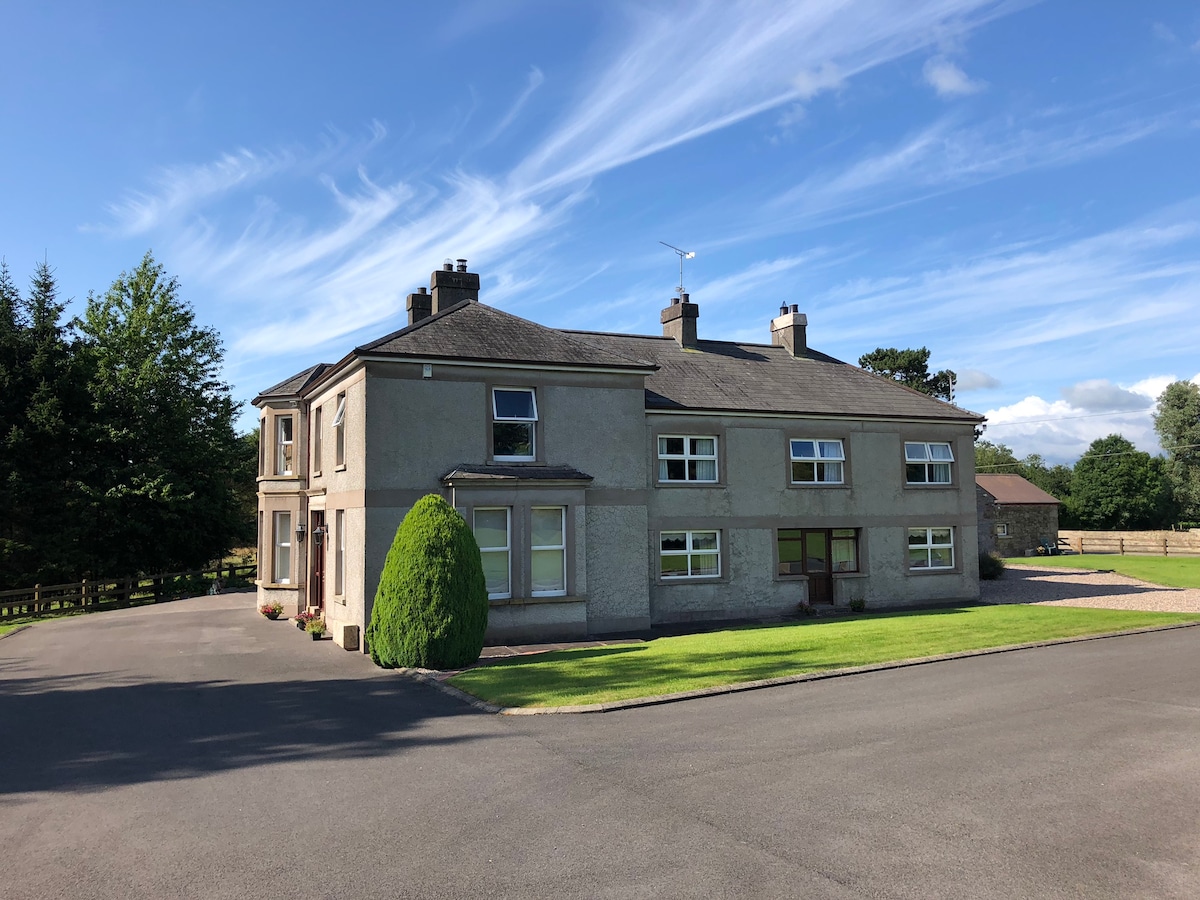 The exterior of Derryvree House is showcased, featuring a two-story structure with large windows and a welcoming gravel driveway. Lush green lawns and neatly trimmed hedges surround the home, while a clear blue sky with wispy clouds provides a serene backdrop.