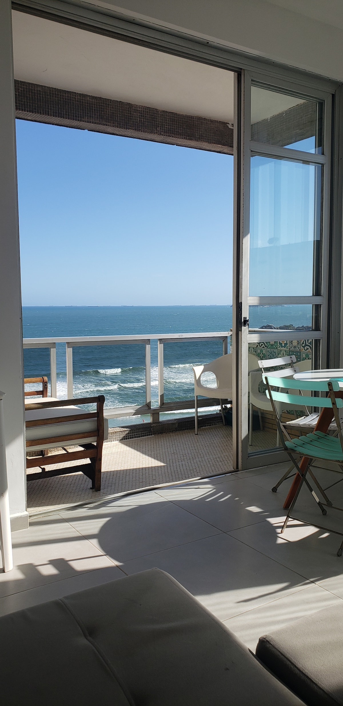 A bright living area opens to a balcony with stunning views of the ocean. The space features a dining table and chairs, allowing for relaxed gatherings. Large glass doors invite natural light and create a seamless connection with the outdoor scenery.