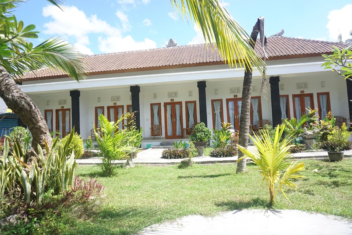 The image captures a row of rooms set in a tropical garden. Large wooden doors with glass panels open to lush greenery and well-kept plants. A concrete path leads to the entrance, surrounded by palm trees and flowering shrubs under a bright blue sky.