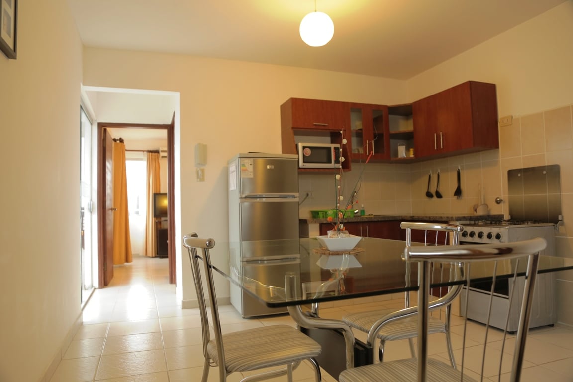 The kitchen area features modern appliances, including a refrigerator and stove, complemented by wooden cabinetry. A glass dining table with metal chairs is positioned centrally, while warm light from a ceiling fixture enhances the inviting atmosphere. A doorway to the living area is visible in the background.