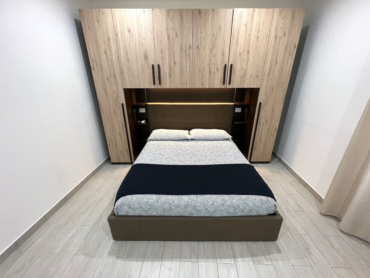 A spacious bedroom features a comfortable bed with a dark blue duvet. The bed is integrated into a modern wooden wall unit that includes built-in shelves on either side. Soft lighting accentuates the space, while neutral-colored walls enhance the bright atmosphere.