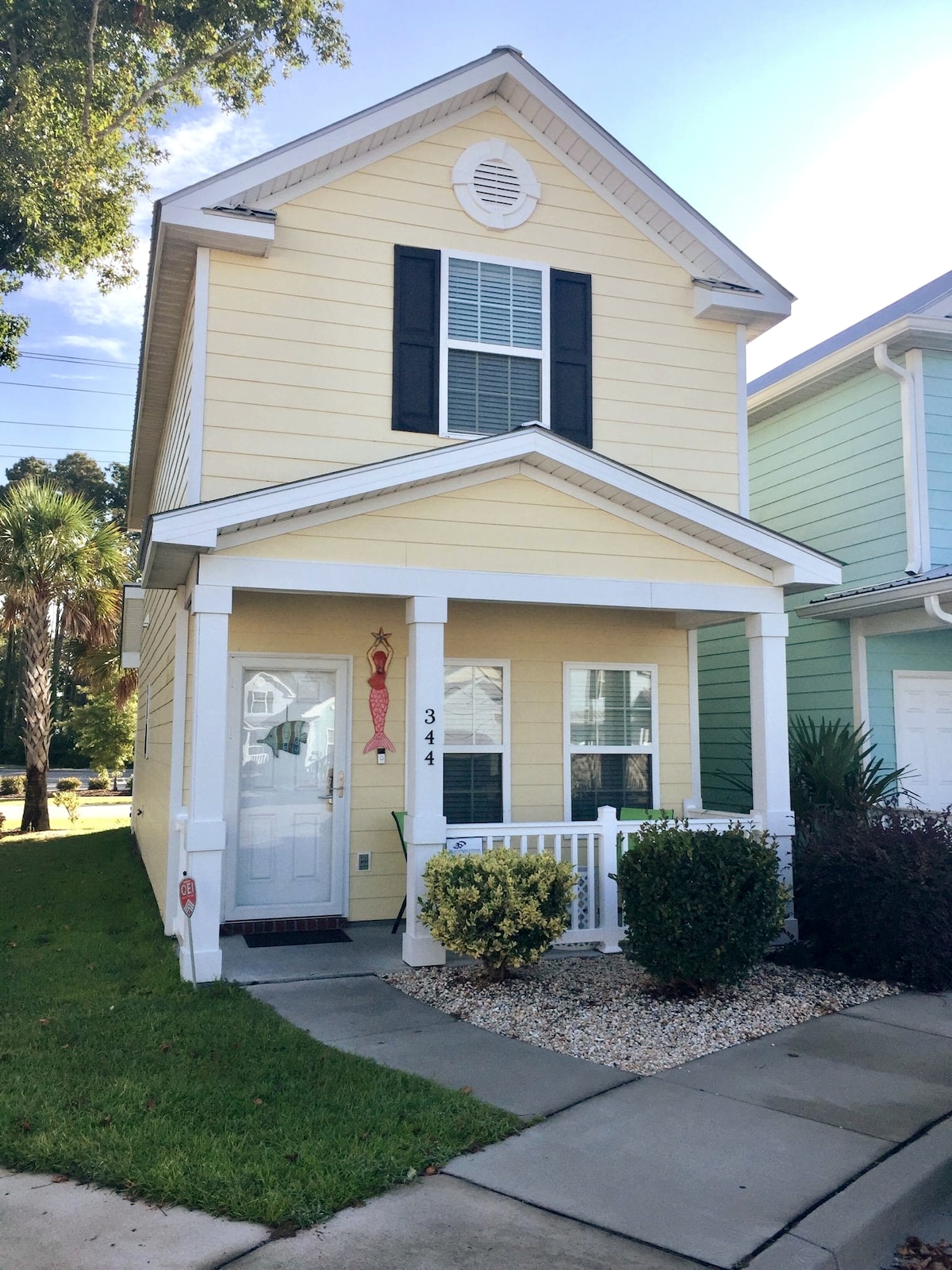 A charming yellow cottage is presented, featuring white trim and a welcoming porch. A decorative red element hangs near the front door. Neatly landscaped with green bushes and small shrubs, the exterior is complemented by a paved pathway and nearby palm trees, enhancing the inviting atmosphere.