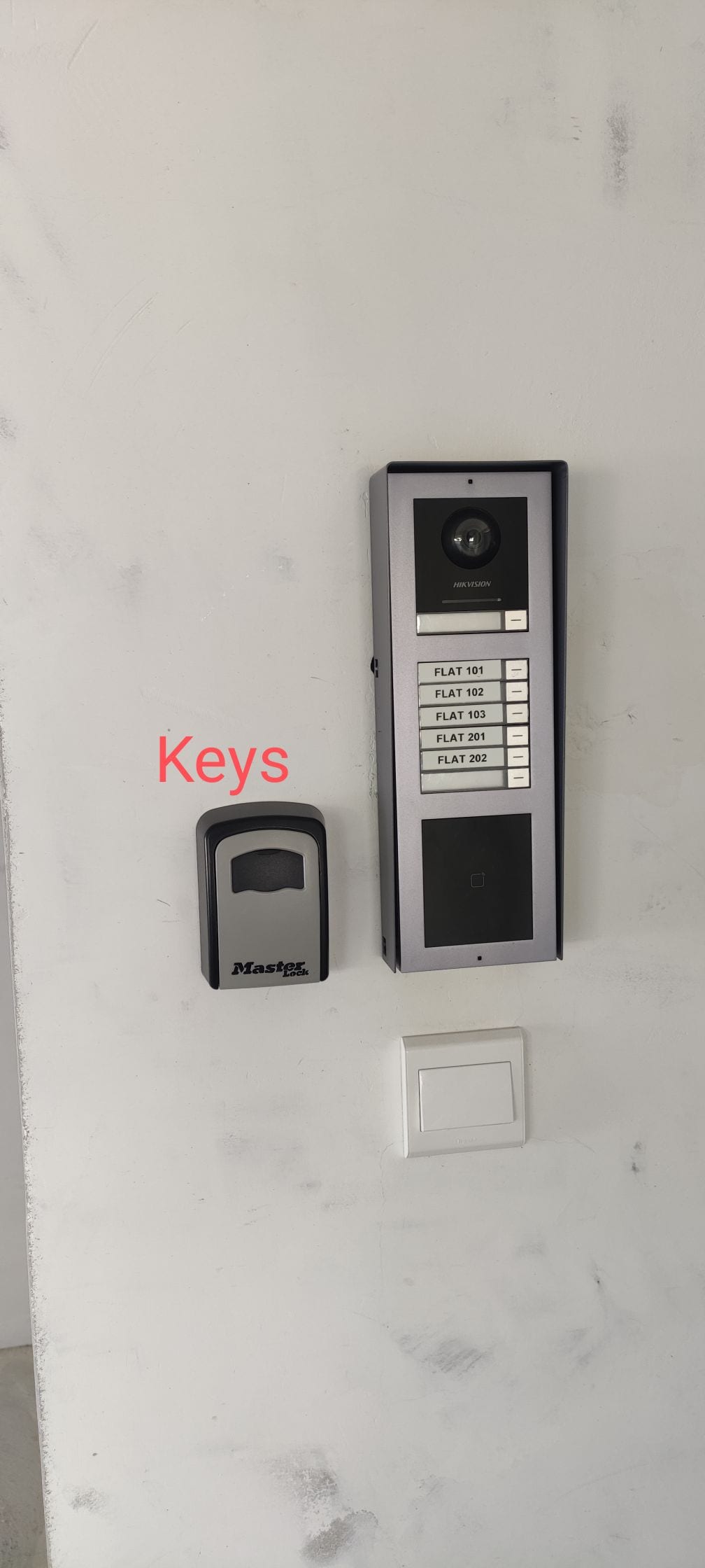 A wall-mounted intercom system with a video camera and numbered buttons is shown beside a key access panel. A white switch is located below the intercom, highlighting the building's secure entry features.