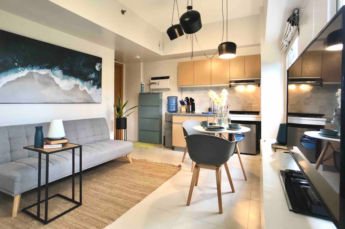 A well-appointed living area is observed with a sleek grey sofa and minimalist coffee table. A dining set with modern chairs features a vase of flowers. The kitchen showcases contemporary cabinetry and appliances, complemented by a large artwork depicting a tranquil beach scene.