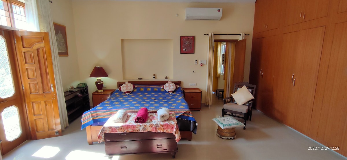 The spacious bedroom features a large bed with a colorful bedspread, complemented by wooden furniture including a bedside table and a rocking chair. Ample natural light enters through a window, enhancing the room's airy feel. Storage cabinets line the wall, providing practical space.