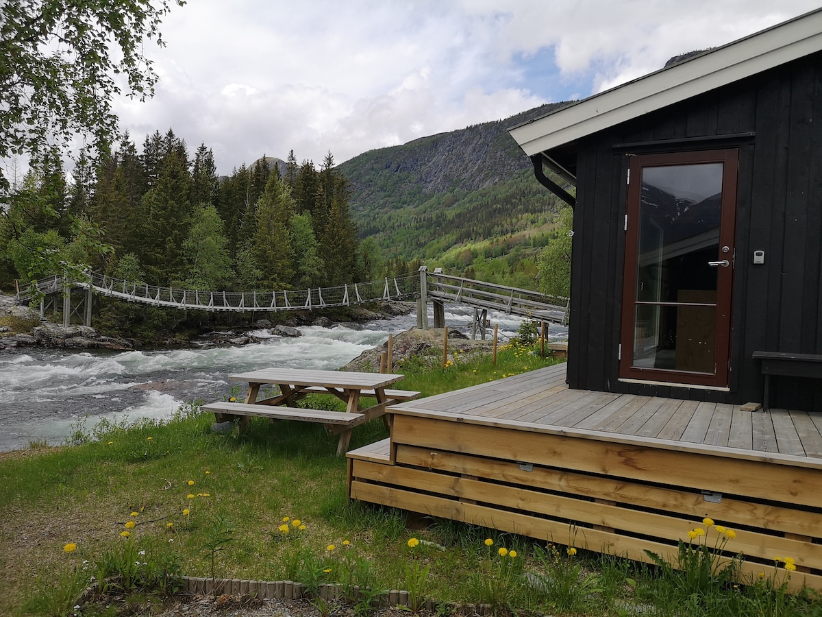A wooden deck extends from the cabin, featuring seating options surrounded by grass and dandelions. A suspension bridge crosses the flowing river, framed by lush green trees and mountains in the distance under a partly cloudy sky.