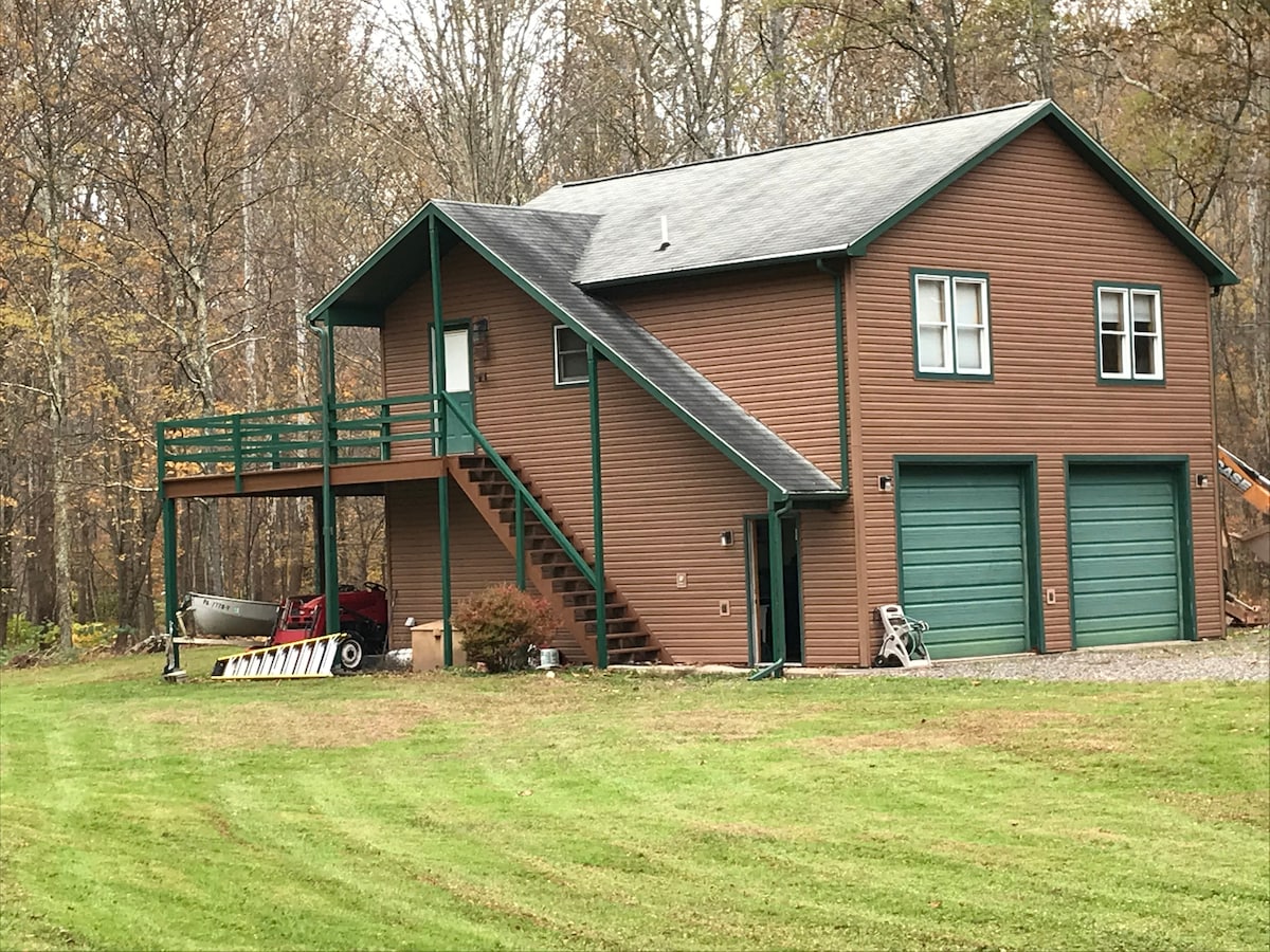 A two-story cabin is set within a wooded area, showcasing a brown exterior with green trim. The upper level features a deck with stairs leading down to the yard. Two garage doors are visible at ground level, accompanied by outdoor equipment and a boat.