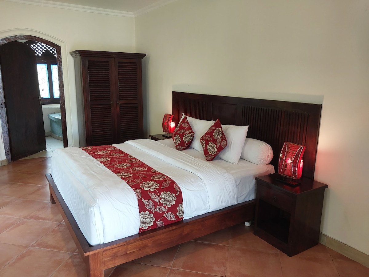 A spacious sleeping area features a large bed with a decorative red and white floral bed runner. Two bedside tables with elegant lamps flank the bed. A wooden wardrobe is positioned against the wall, and an accessible area leads to a bathroom in the background.