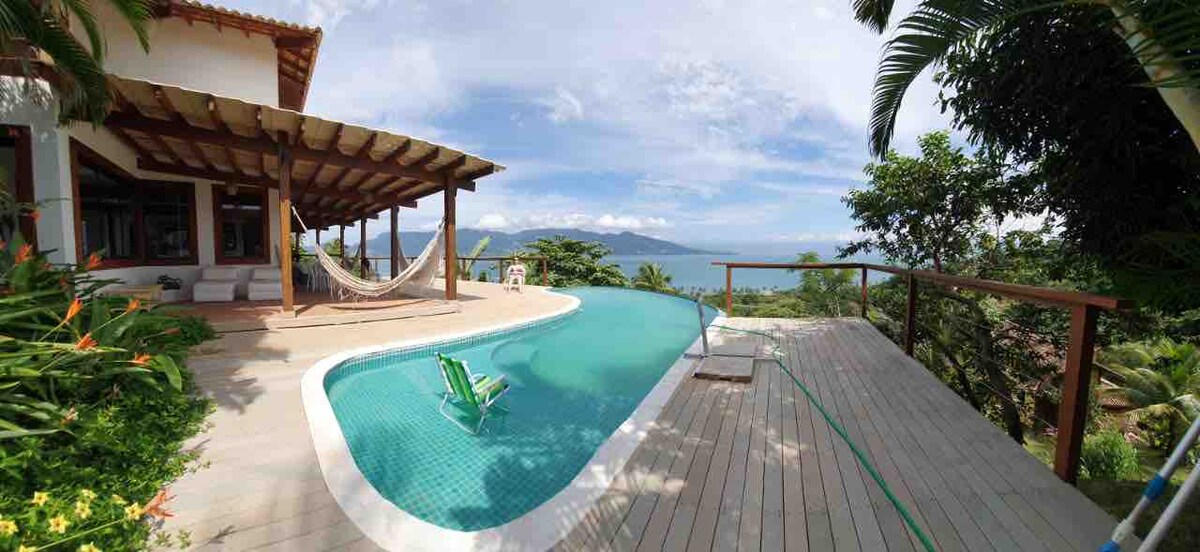 The outdoor area features a spacious pool with a view of the ocean, surrounded by tropical greenery. A wooden deck complements the pool, while a hammock is suspended nearby, inviting relaxation. The sky above is bright, offering a serene backdrop to the scene.