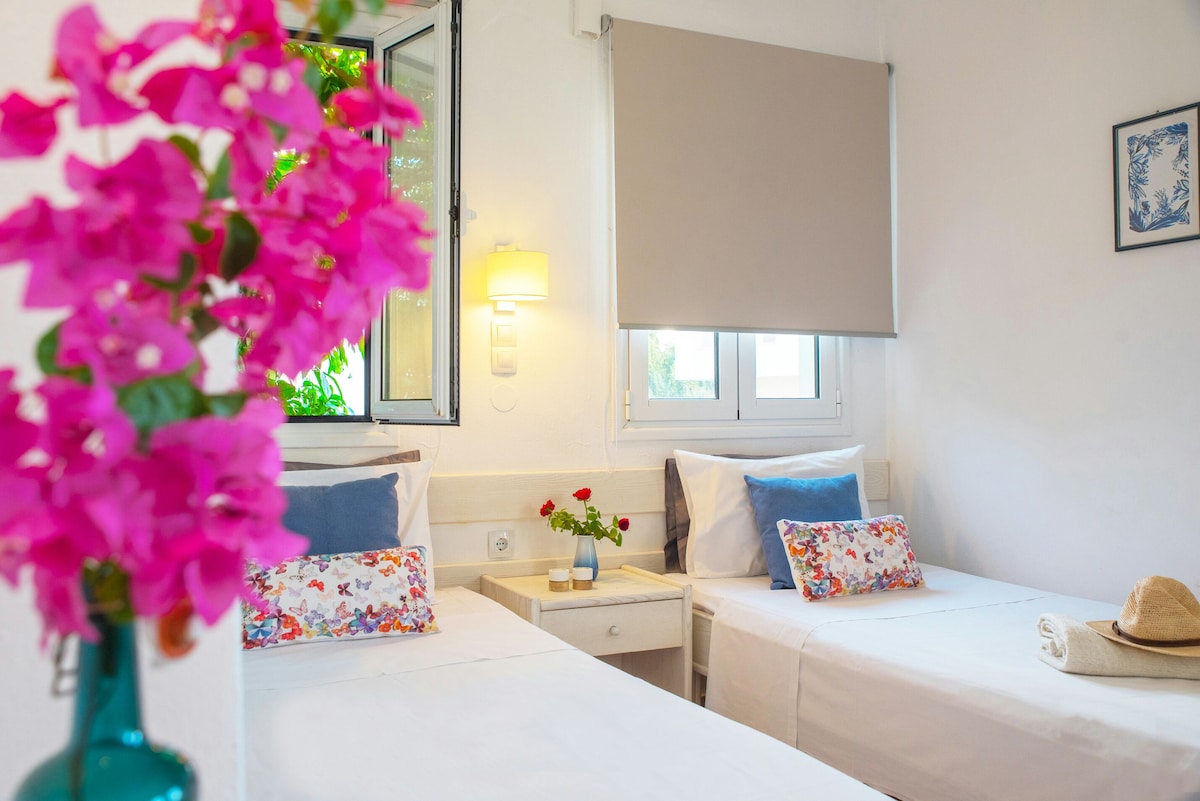Two twin beds are positioned against a bright white wall, each adorned with decorative cushions. A small bedside table sits between them, complemented by a lamp. Natural light enters through the windows, framed by light gray shades, while vibrant pink flowers add a touch of color.
