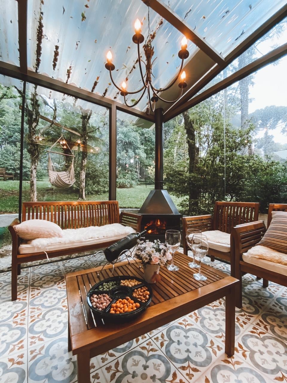 A glass-enclosed seating area features wooden benches and a table, accompanied by a stylish chandelier. A cozy fireplace is positioned centrally, with a bowl of snacks on the table and views of a lush green outdoor space visible through the transparent walls.