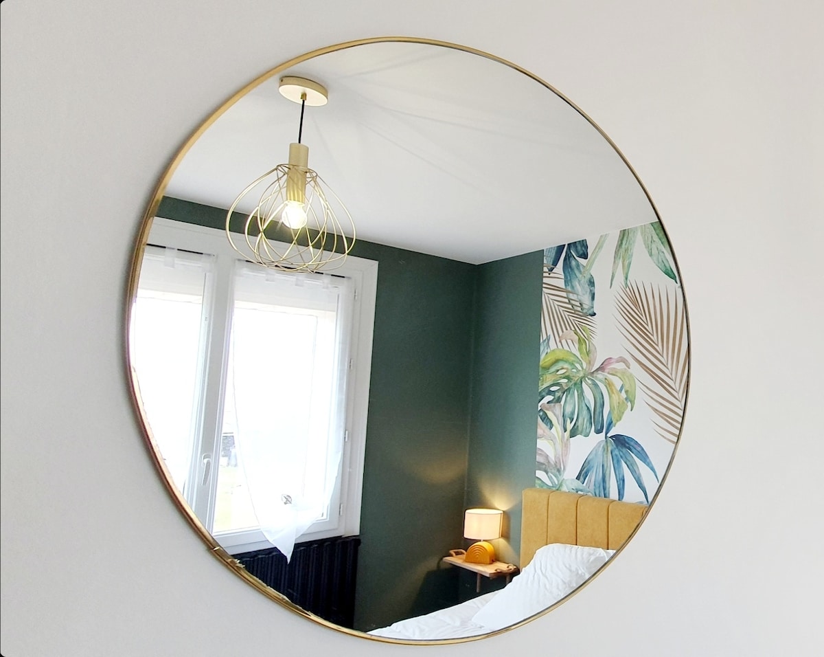 A round mirror framed in gold reflects the ceiling light and contributes to the room's design. Behind it, a patterned wall featuring tropical leaves complements the muted green walls. Two windows allow natural light, while a bedside lamp rests next to a neatly made bed.