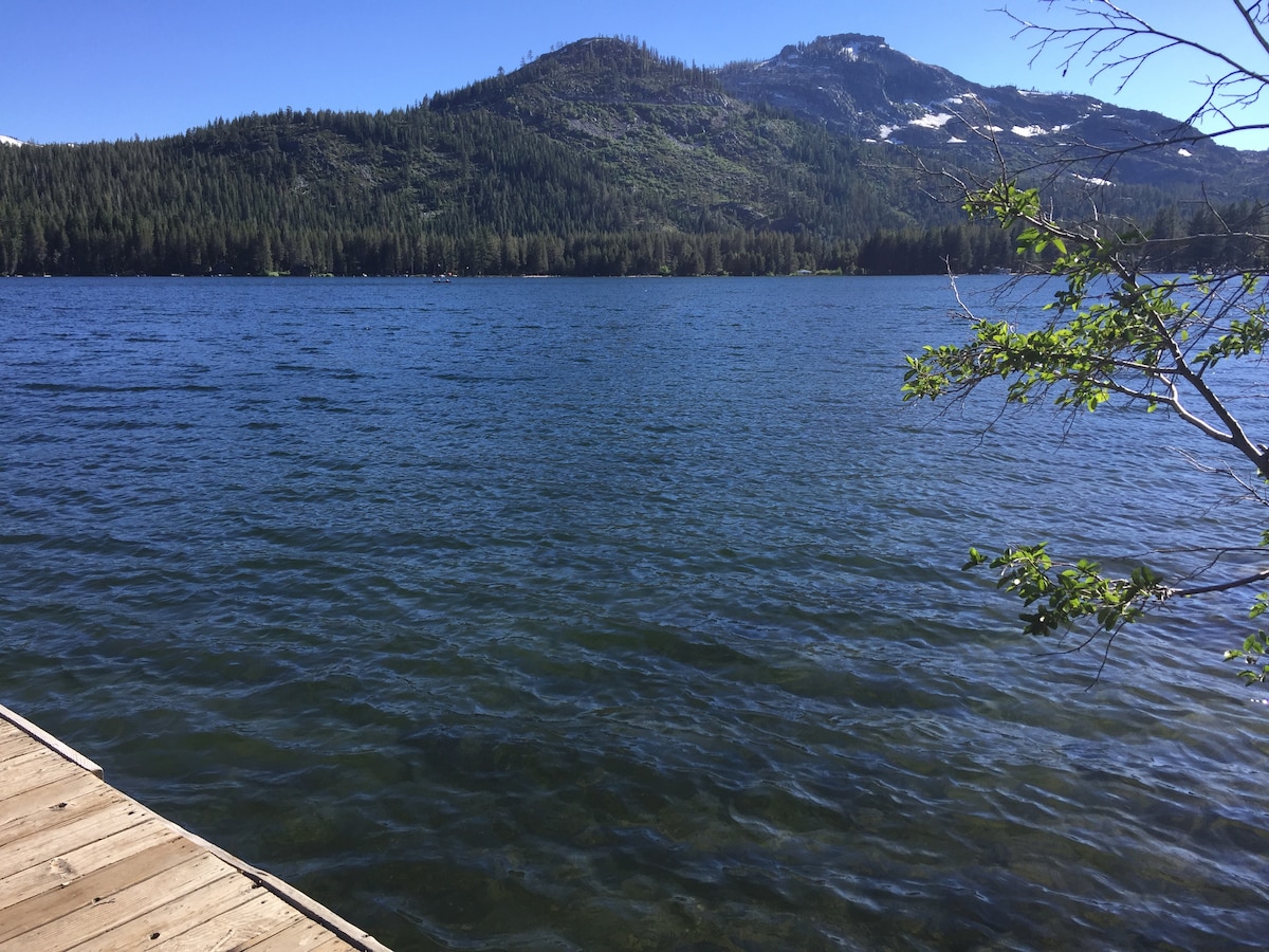 A clear view of Donner Lake is presented, with gentle waves reflecting the sunlight. Lush green trees line the shore, and a mountainous backdrop creates a serene atmosphere. The wooden dock extends into the water, inviting guests to enjoy the beauty of the lake.