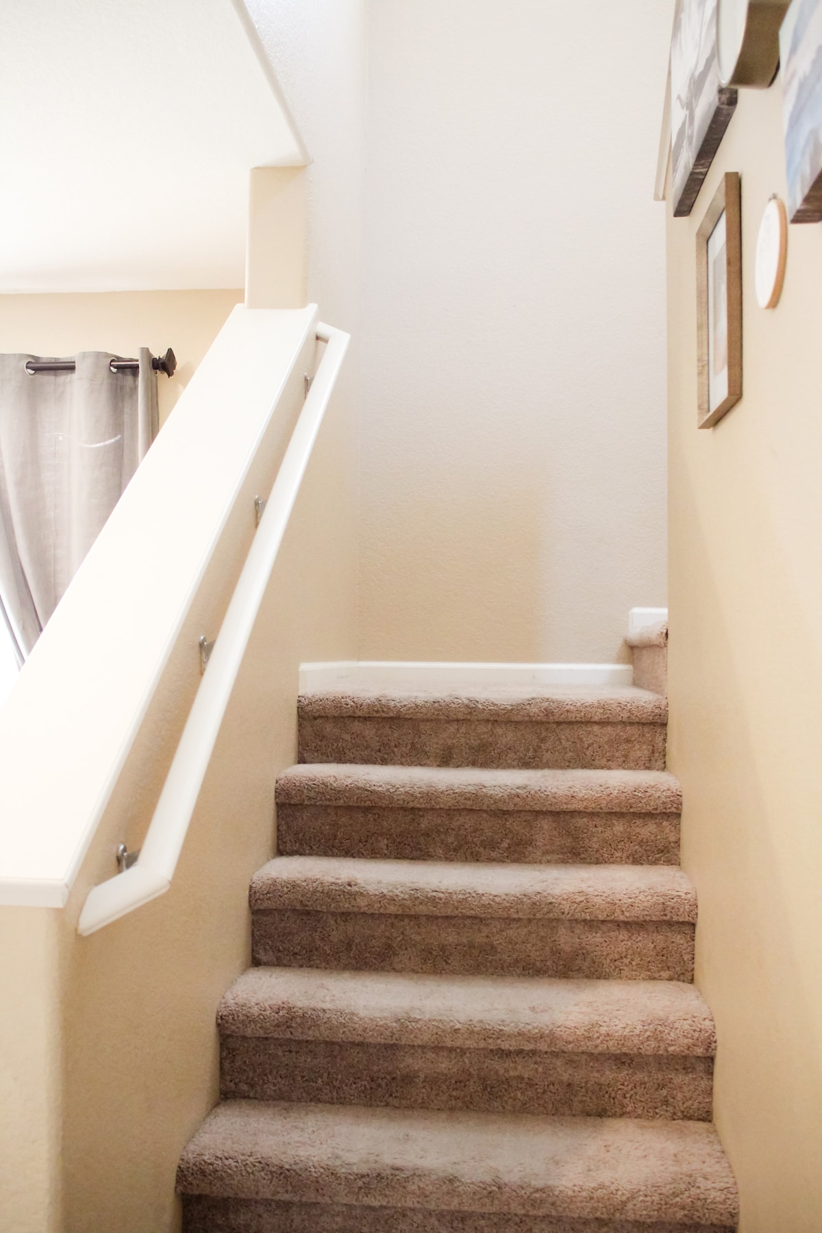 A staircase with beige carpeting leads to the upper level of the home. Walls are painted in a light tone, and a handrail is positioned on the right. Framed photographs are displayed along the wall near the top of the stairs.