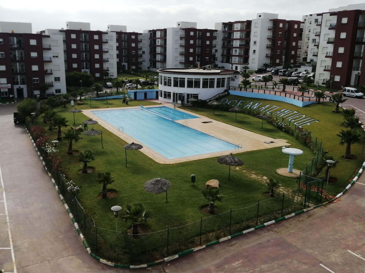 A large outdoor swimming pool is surrounded by manicured lawns and palm trees. Shade is provided by several thatched umbrellas set along the pool's edge. The scene is complemented by a multi-story building in the background, creating a peaceful atmosphere.