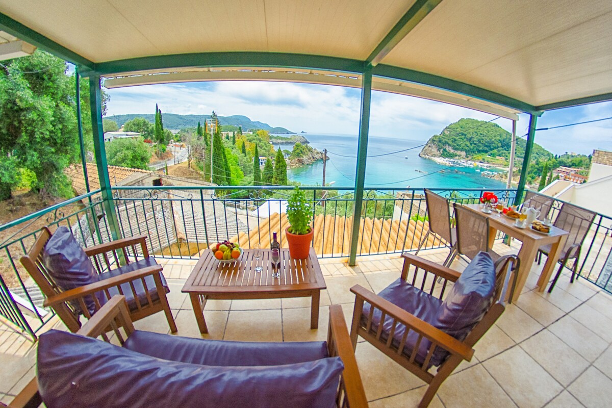 A spacious terrace features wooden furniture, including armchairs and a table set for outdoor dining. A vibrant view of the coastline and sea is visible, framed by lush greenery and distant hills. The setting creates an inviting space for relaxation and enjoyment of the scenery.