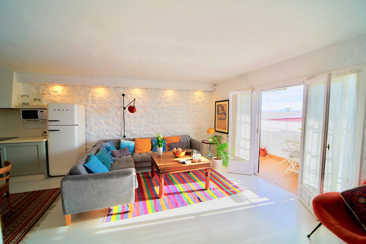 A spacious living area features a large corner sofa adorned with colorful cushions. A vintage coffee table sits in the center, surrounded by a vibrant area rug. Light flows through French doors leading to a balcony, with a small dining set visible outside.