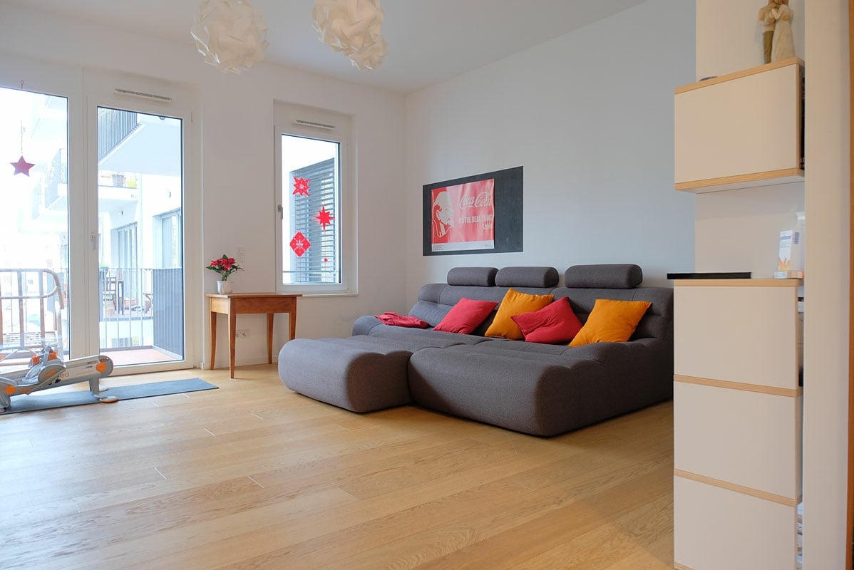 A spacious living area features a large gray couch adorned with colorful cushions. Light wooden flooring complements the bright atmosphere, while floor-to-ceiling windows allow natural light to fill the room. A decorative wall art piece adds a pop of color, enhancing the overall design.