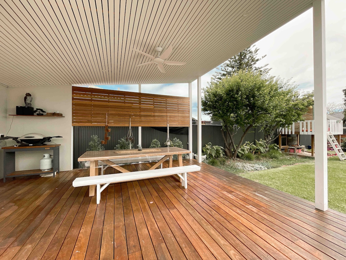 An outdoor deck area features a large wooden table surrounded by benches, providing a space for dining. A ceiling fan allows for airflow under the covered patio, while a garden and play structure are visible in the background.
