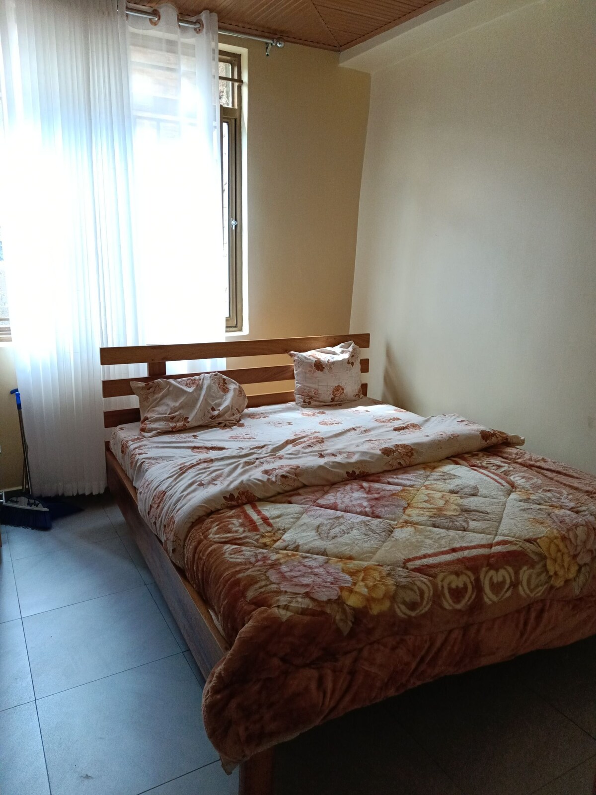 A compact sleeping area features a wooden bed with a decorative quilt. Pillows are arranged at the head of the bed. Natural light enters through a large window dressed with sheer curtains, illuminating the room's neutral walls and tiled floor.