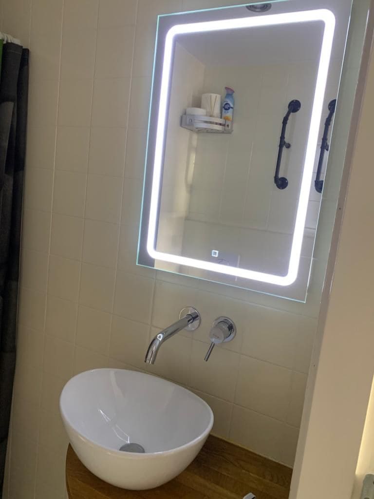 A modern bathroom sink area is featured, showcasing a round, white vessel sink positioned atop a wooden countertop. Above the sink, a backlit mirror is mounted on the wall, while toiletries are neatly organized on a small shelf. The overall aesthetic is clean and functional.