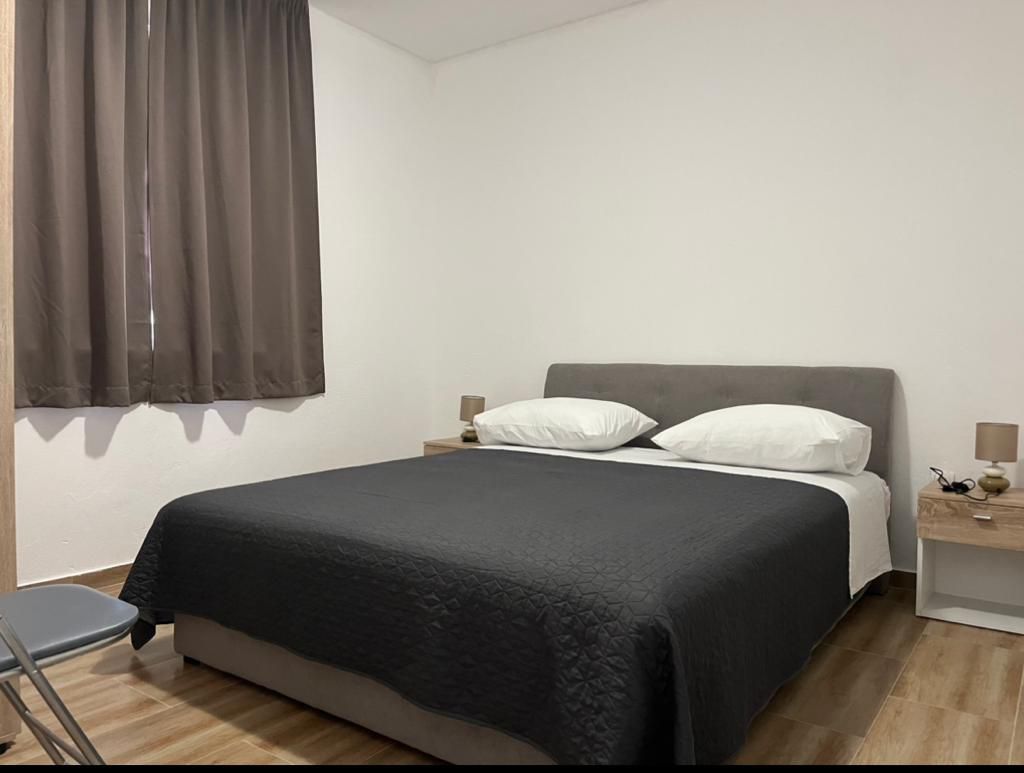 A cozy bedroom features a spacious bed with dark bedding and white pillows, flanked by bedside tables with lamps. Natural light is partially filtered through floor-length curtains, while warm wooden flooring contributes to a comfortable environment.