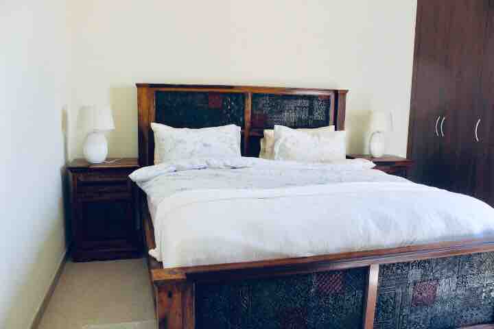 A spacious bedroom is presented with a large wooden bed featuring an intricately designed headboard. Two bedside tables with lamps are placed on either side of the bed. A sizable wardrobe is visible against one wall, contributing to the room's organized appearance.