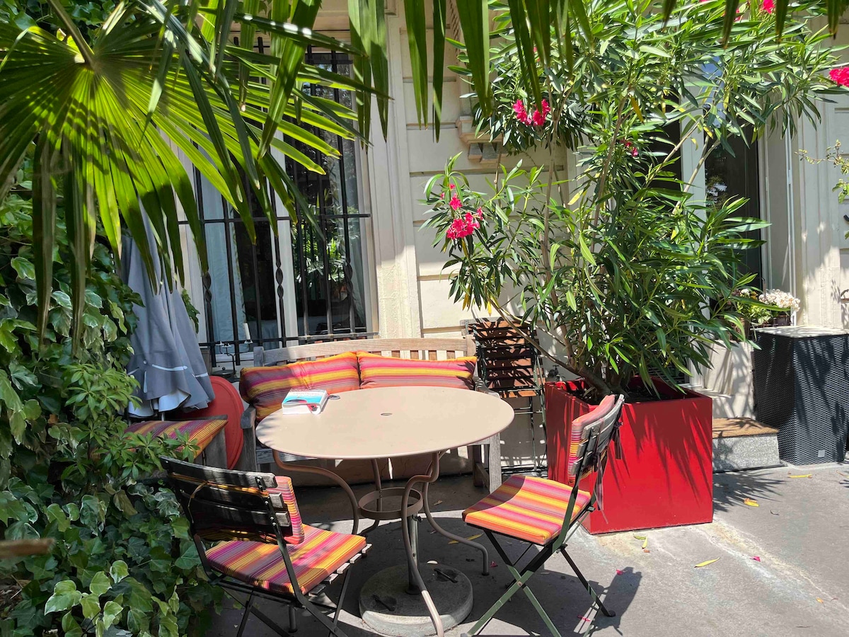 A private garden space features a round table surrounded by colorful striped chairs. Lush greenery and flowering plants enhance the inviting environment. A comfortable seating area with cushions is present, providing a cozy spot for relaxation. Sunlight filters through the foliage, creating a pleasant atmosphere.