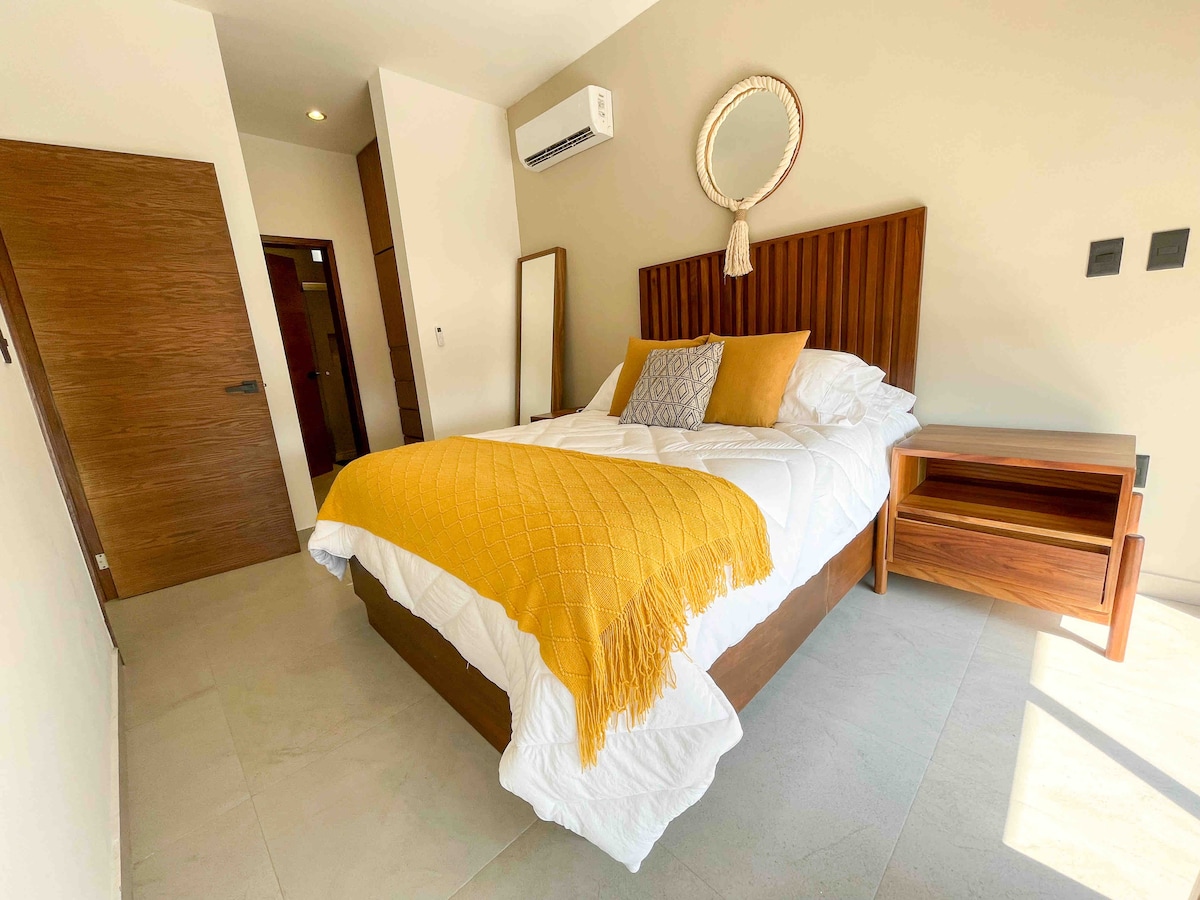 A bright bedroom features a spacious bed with a yellow knitted throw and decorative pillows. A wood-framed mirror and bedside table complement the room's design. Natural light fills the space, highlighting the neutral wall tones and subtle decor elements.