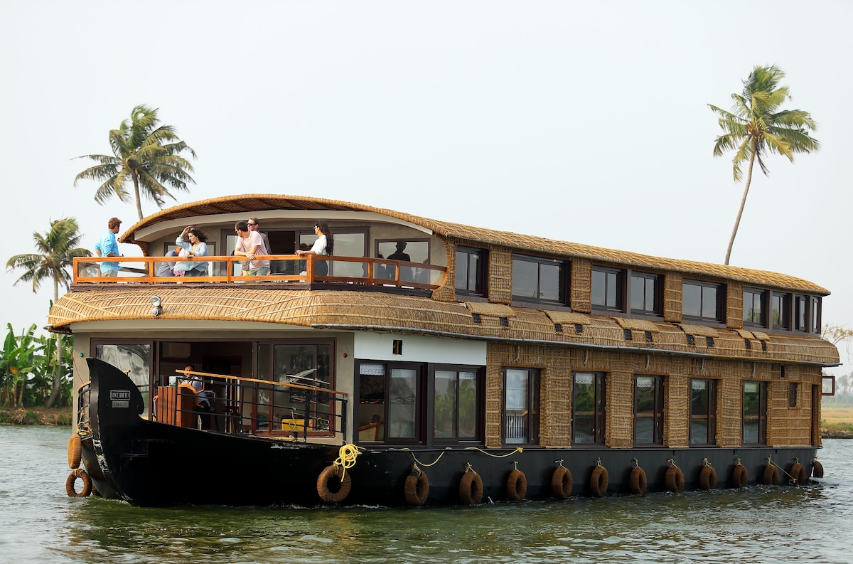 A large, two-story houseboat floats gently on the water. The structure features a thatched roof and spacious balconies, while numerous windows offer views of the surroundings. Palm trees line the background, reflecting the tranquil atmosphere of the backwaters.