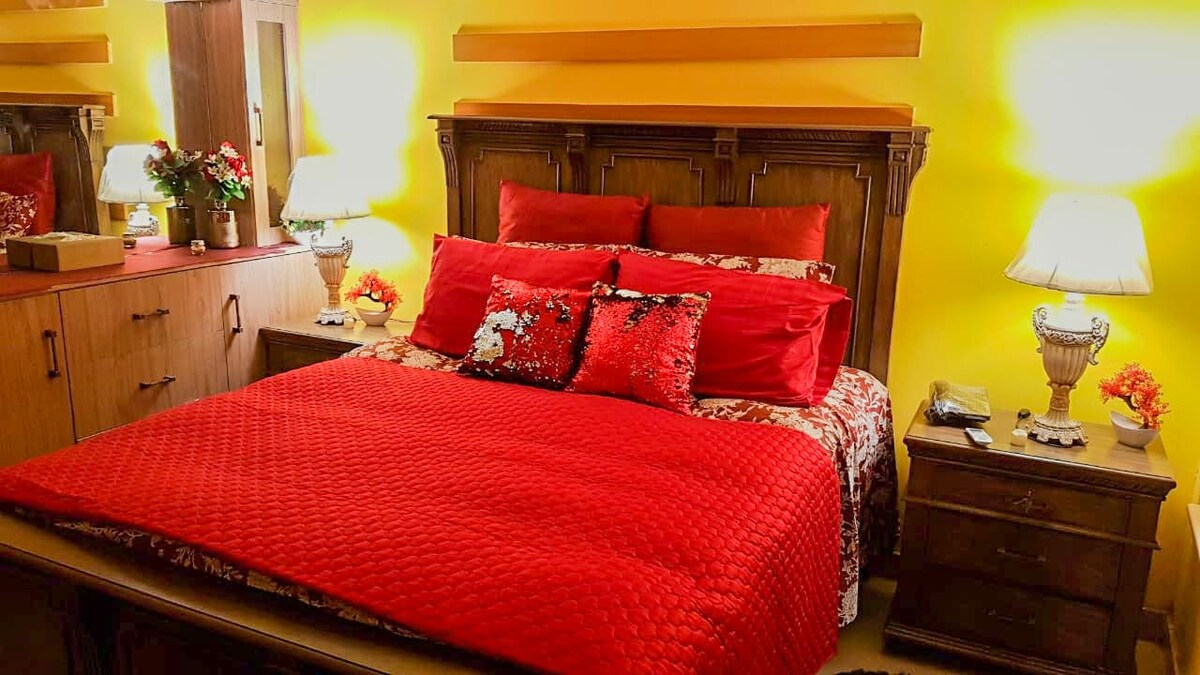 A spacious bed is arranged with red and white bedding, complemented by an assortment of decorative pillows. Nightstands with lamps are positioned on either side, and a wooden dresser is visible, creating a cozy and vibrant sleeping area with warm yellow walls.
