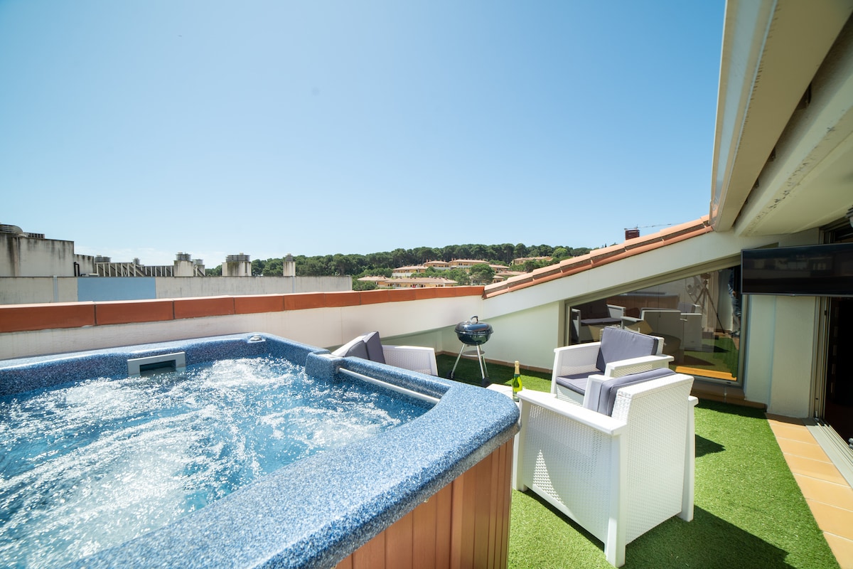 A private terrace is featured, showcasing a jacuzzi beside comfortable outdoor seating. A portable barbecue is also present, with bright blue skies above and greenery visible in the distance, creating a relaxing outdoor space.