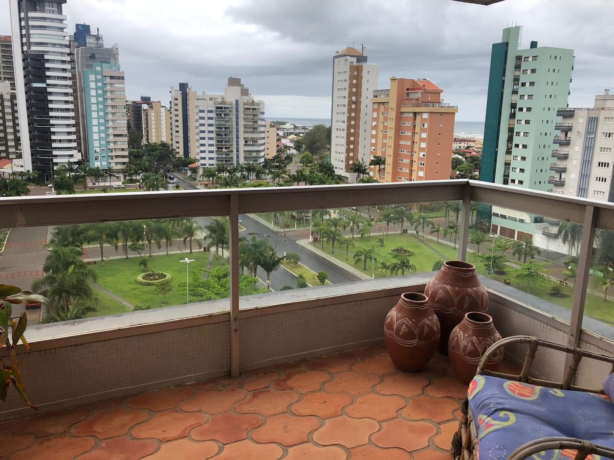 An outdoor balcony space showcases clay pots alongside colorful seating, offering a view of lush greenery and city buildings. The landscape includes neatly arranged parks and a glimpse of the ocean in the distance, framed by a cloudy sky.