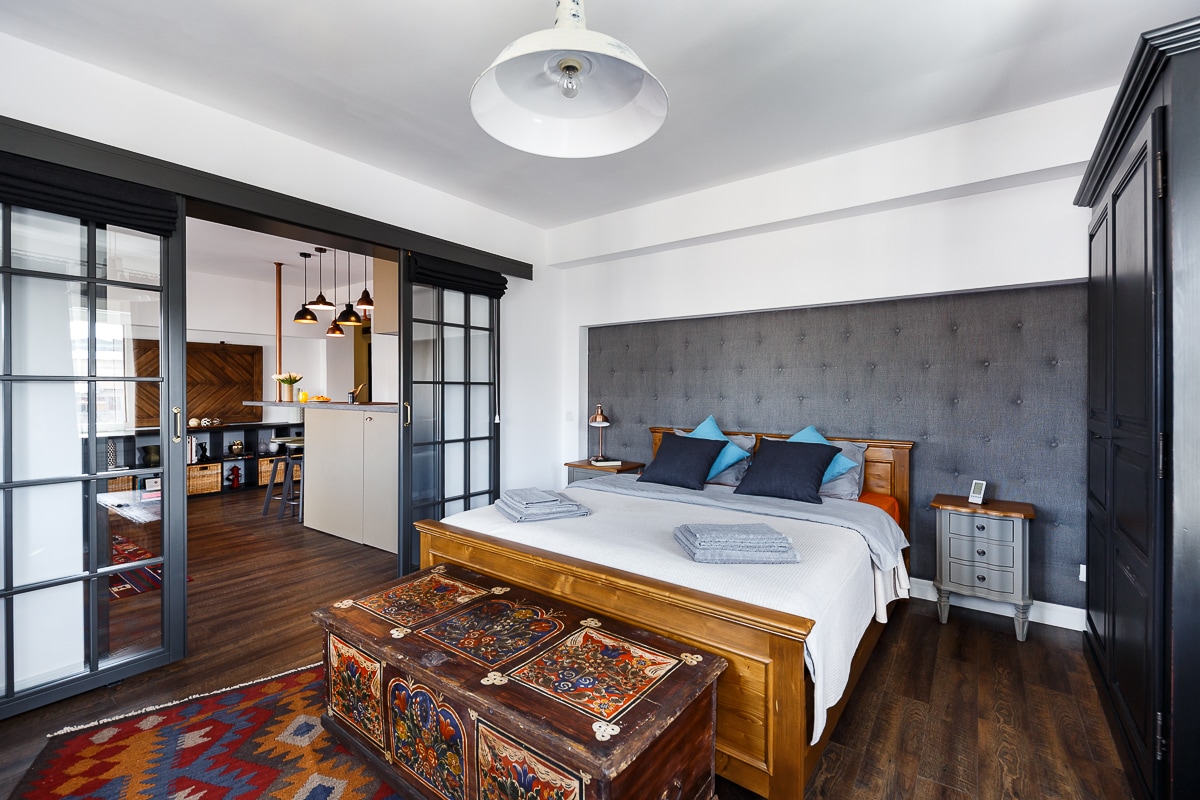 A spacious bedroom features a large designer bed with decorative pillows. Traditional Romanian furniture, including a polychrome dowry chest and a dark wooden cabinet, adds character. Large windows allow ample natural light, and the room is designed with warm wooden tones and eclectic decor.