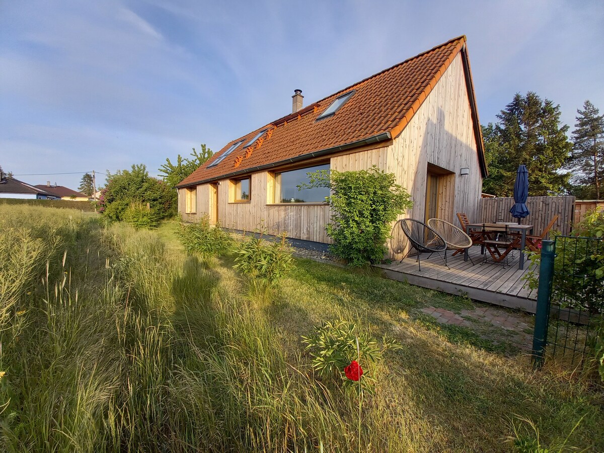 A modern wooden house features a sloped roof and large windows, allowing ample natural light. Outside, a deck with a dining area is surrounded by lush green grass and landscaping. A lone flower adds a touch of color amid the serene outdoor setting.