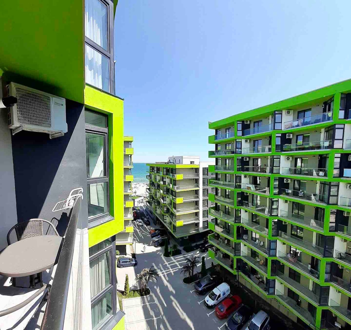 A view from a balcony showcases the vibrant green exterior of the Alezzi Beach Resort complex. Cars are parked below, and glimpses of the sea can be seen in the distance, framed by the modern architectural design of the buildings.
